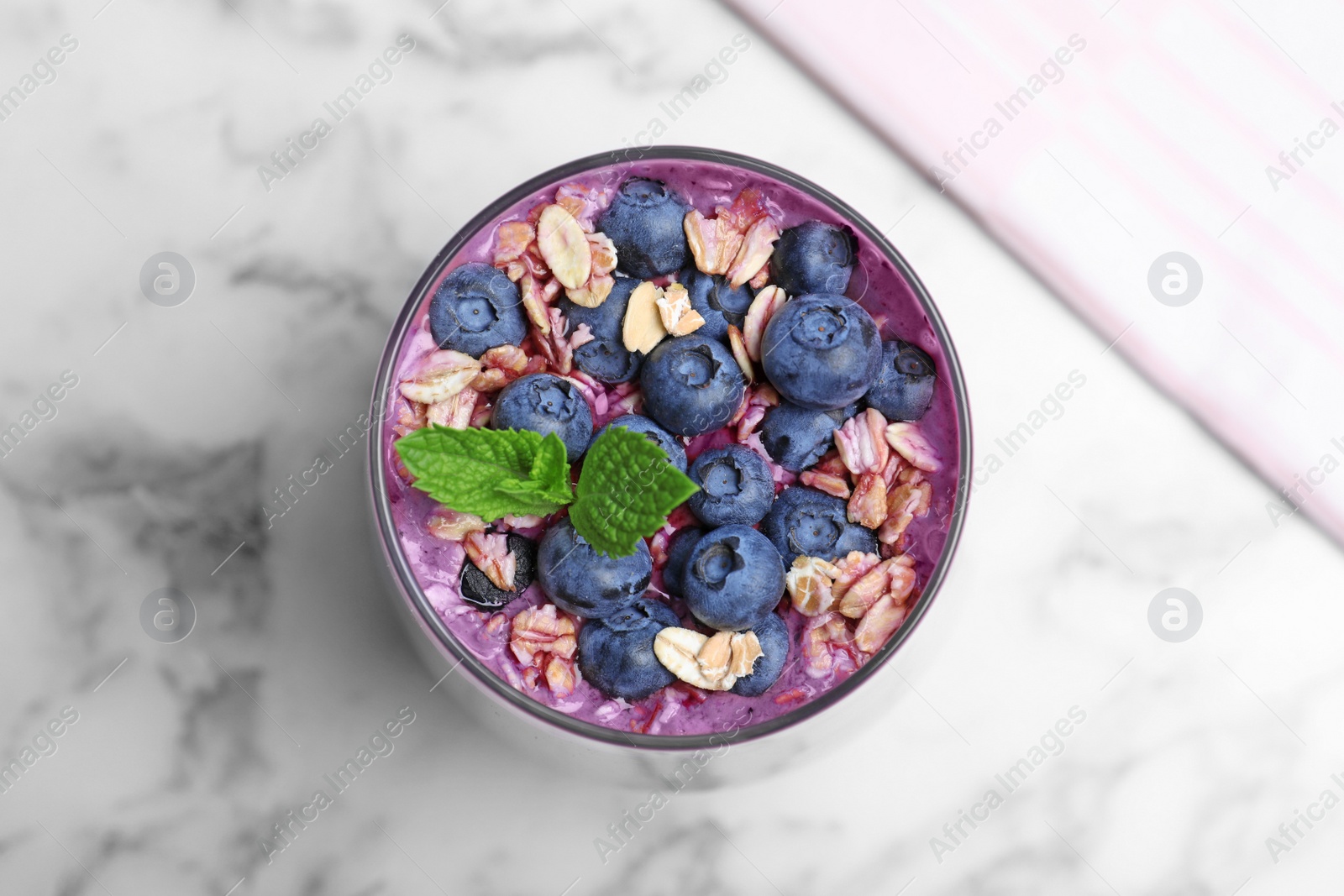 Photo of Glass of delicious blueberry smoothie with granola and mint on white marble table, top view