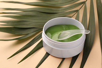 Photo of Jar of under eye patches with spoon and palm leaves on beige background, closeup. Cosmetic product