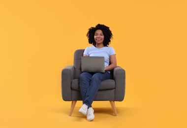 Happy young woman with laptop sitting in armchair against yellow background