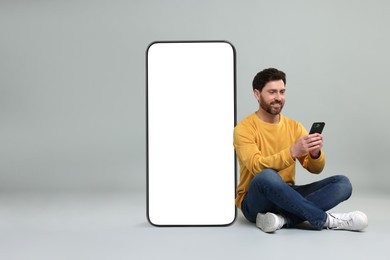 Man with mobile phone sitting near huge device with empty screen on grey background. Mockup for design