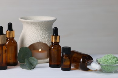 Photo of Different aromatherapy products, burning candle and eucalyptus leaves on white wooden table against light background