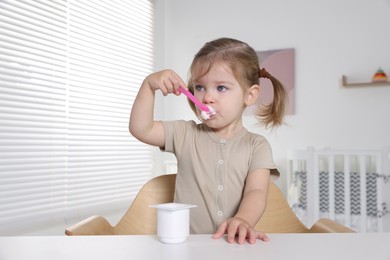Cute little child eating tasty yogurt from plastic cup with spoon at white table indoors