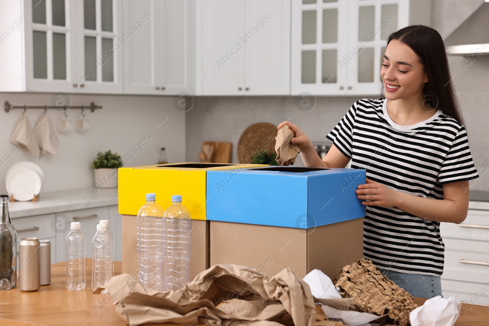 Photo of Garbage sorting. Smiling woman throwing crumpled paper into cardboard box in kitchen
