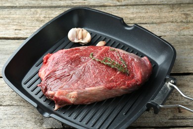 Photo of Grill pan with piece of raw beef meat, garlic and thyme on wooden table