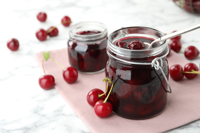 Jars of pickled cherries and fresh fruits on white marble table, closeup