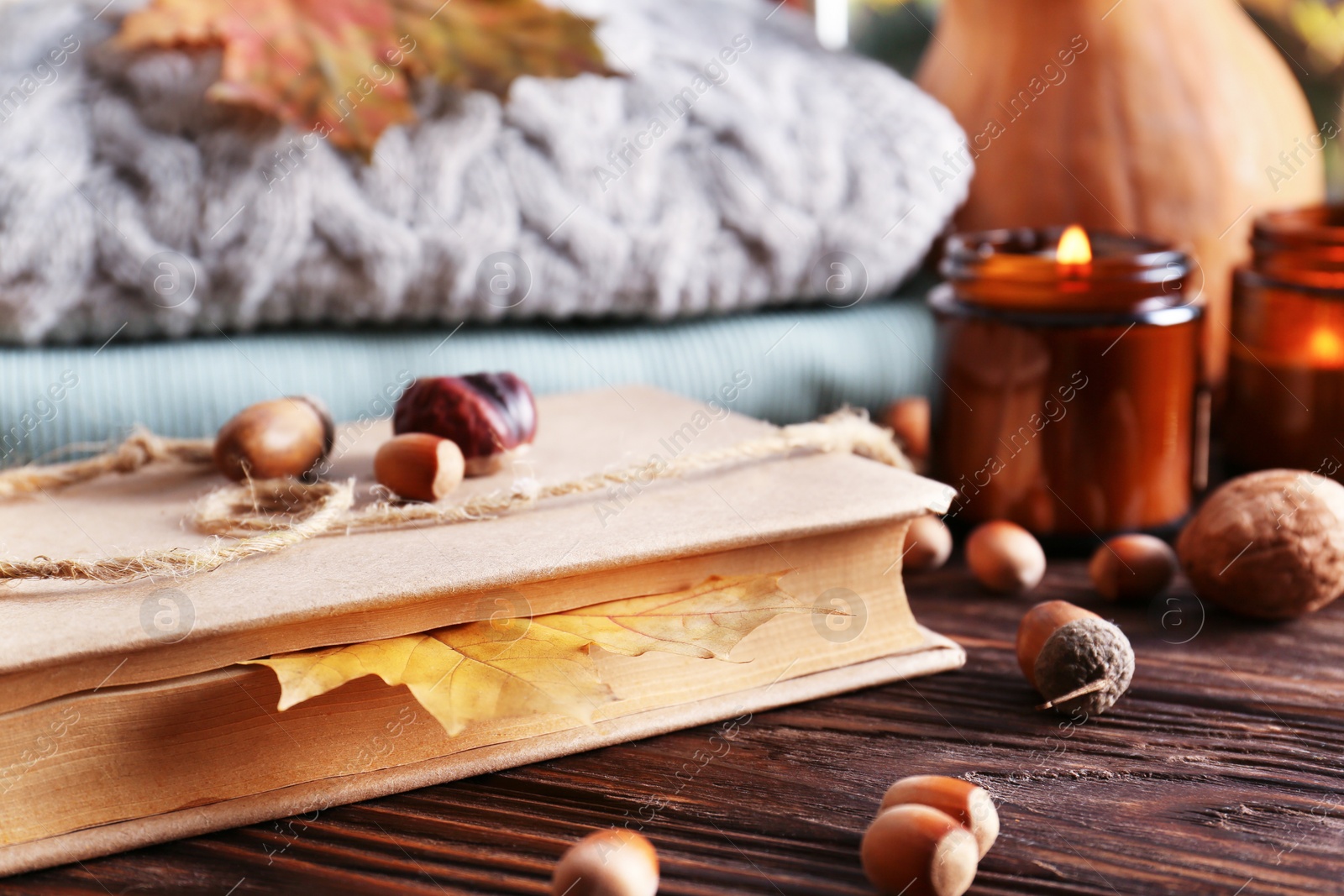 Photo of Book with autumn leaf as bookmark, scented candles and warm sweaters on wooden table, closeup