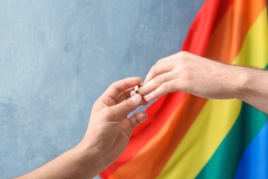 Photo of Young man putting wedding ring on his boyfriend's finger near rainbow flag. Gay marriage