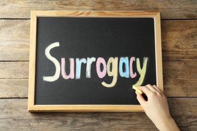 Woman writing word Surrogacy on small blackboard at wooden table, top view