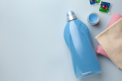 Photo of Bottle of fabric softener, laundry capsules and stacked clean clothes on light background, flat lay. Space for text