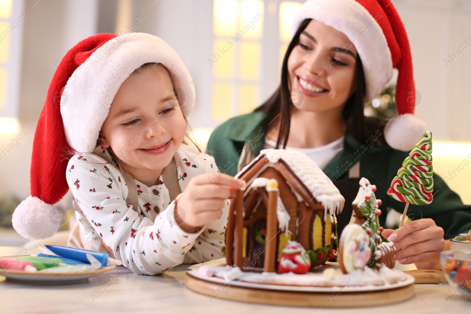 Photo of Mother and daughter decorating gingerbread house at table indoors