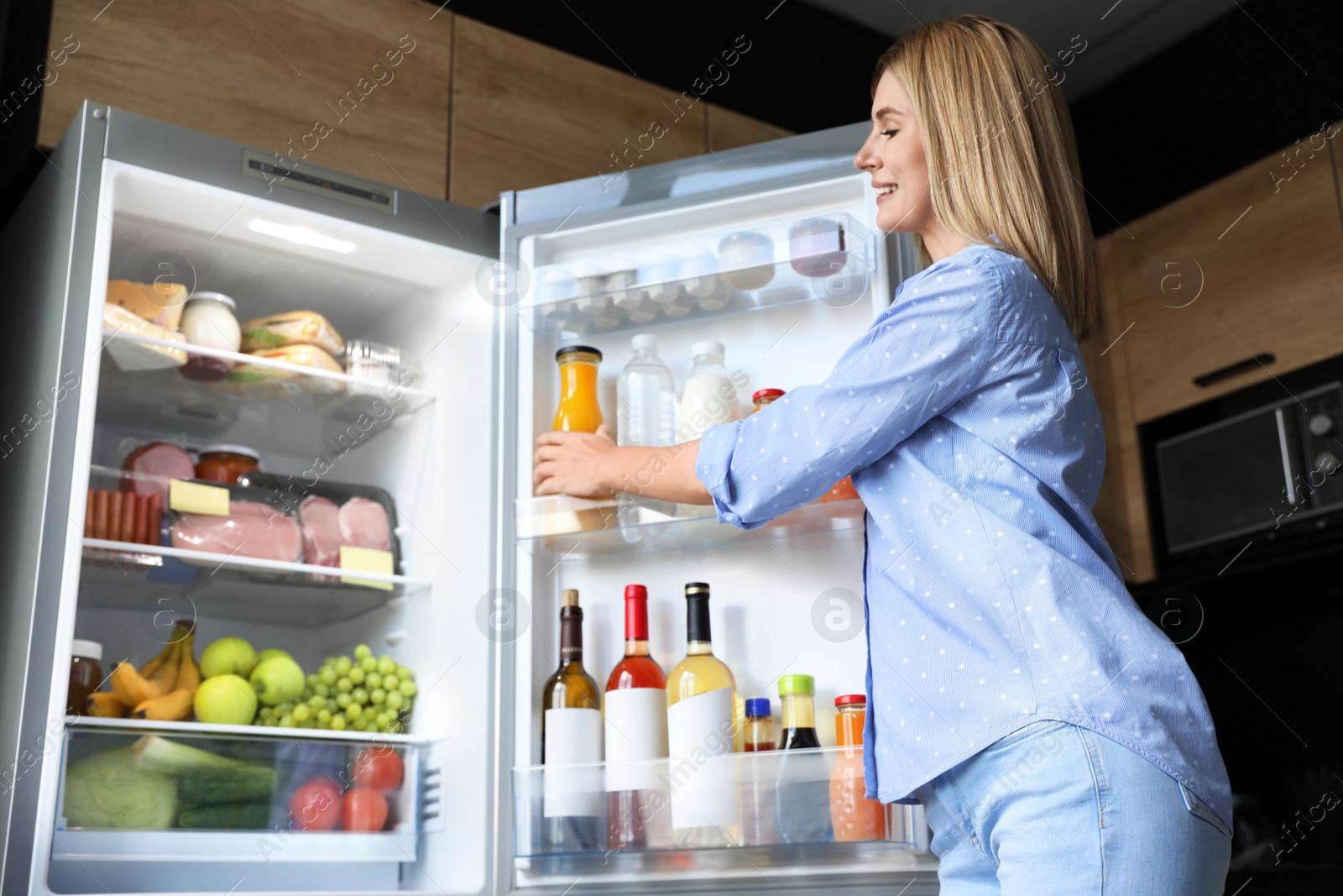 Photo of Woman taking bottle with juice out of refrigerator in kitchen