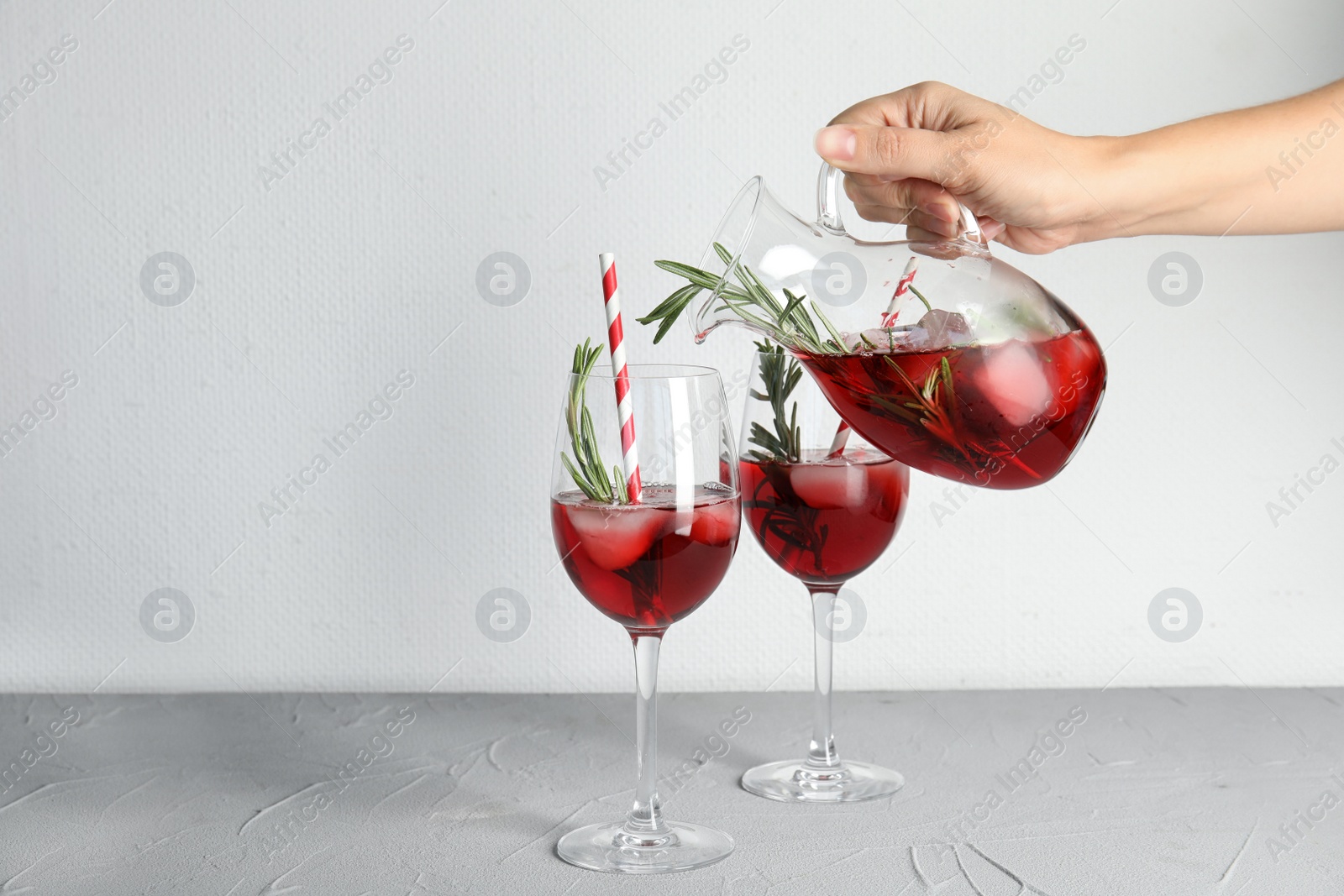 Photo of Woman pouring cranberry cocktail into glass with rosemary on table