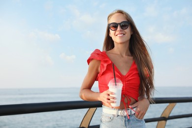 Beautiful woman with tasty milk shake near sea, space for text