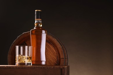 Photo of Whiskey with ice cubes in glass and bottle on wooden table near barrel against dark background, space for text