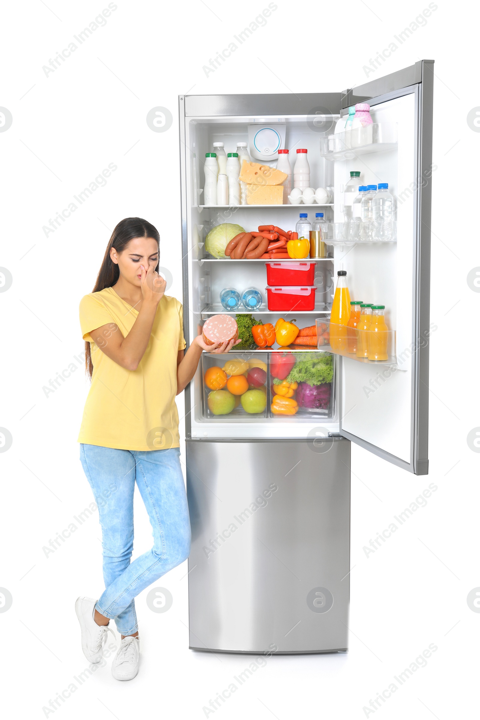 Photo of Young woman with expired sausage near open refrigerator on white background