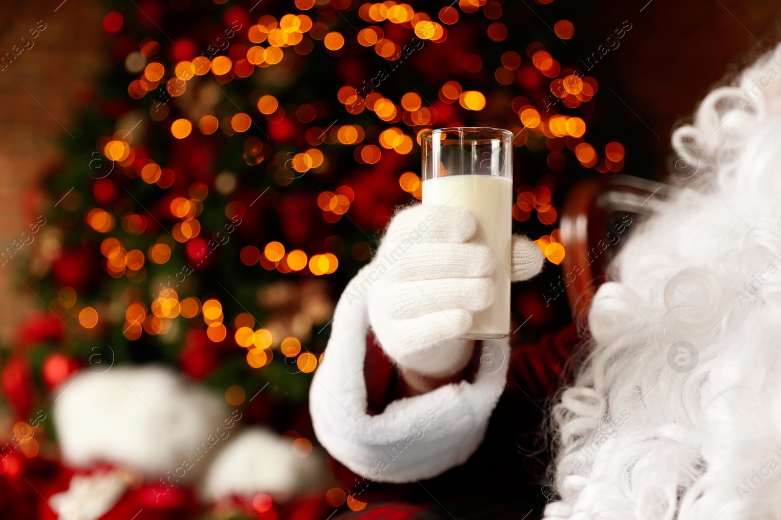 Photo of Santa Claus holding glass of milk against blurred Christmas lights, closeup