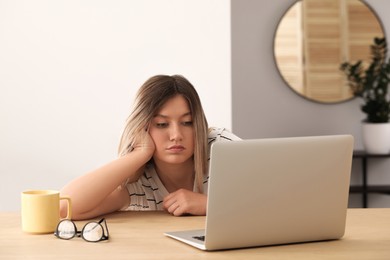 Sleepy young woman with laptop at wooden table indoors