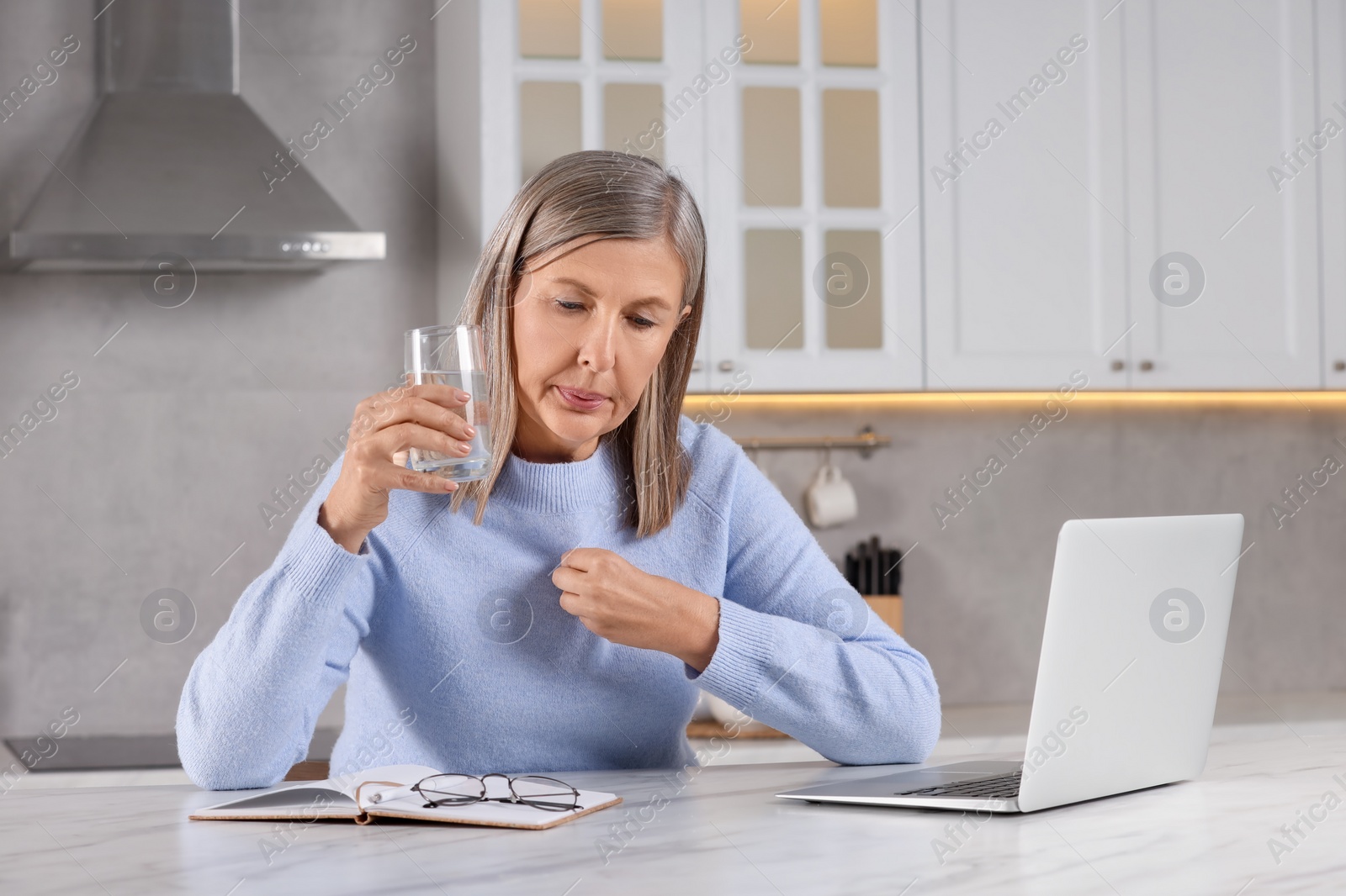 Photo of Menopause. Woman with glass of water suffering from hot flash at table in kitchen