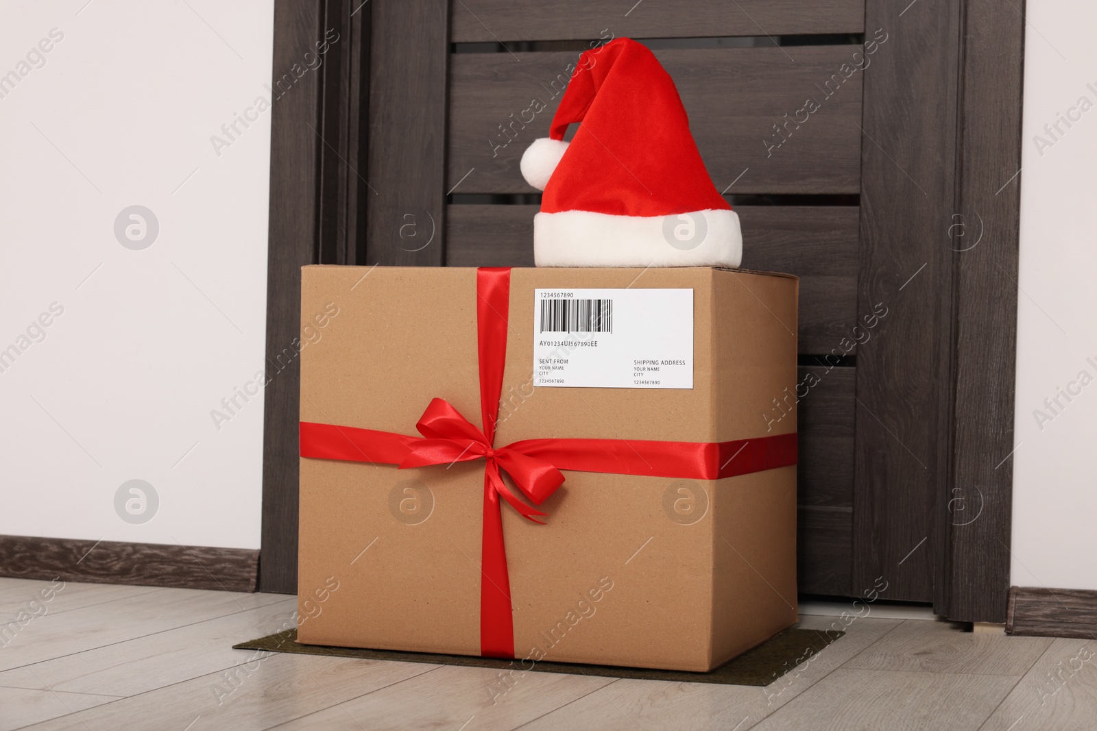 Photo of Christmas gift box and Santa hat near door indoors. Sending present by mail