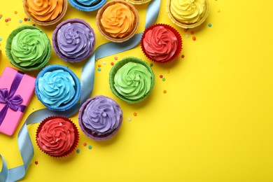 Photo of Delicious birthday cupcakes, gift, ribbon and sprinkles on yellow background, flat lay. Space for text