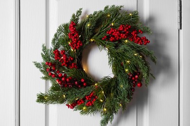 Photo of Beautiful Christmas wreath with red berries and fairy lights hanging on white door