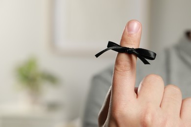 Man showing index finger with black tied bow as reminder on blurred background, closeup. Space for text