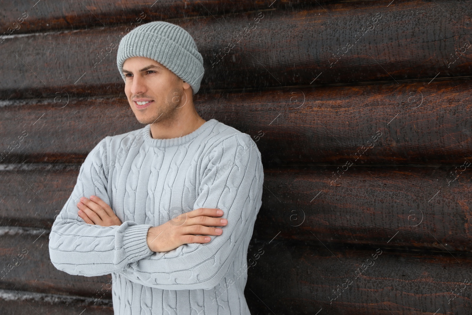 Photo of Handsome man wearing warm sweater and hat near wooden wall, space for text. Winter season