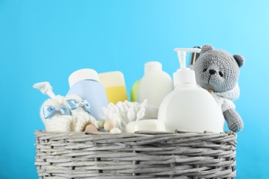 Wicker basket full of different baby cosmetic products, bathing accessories and toy on light blue background, closeup