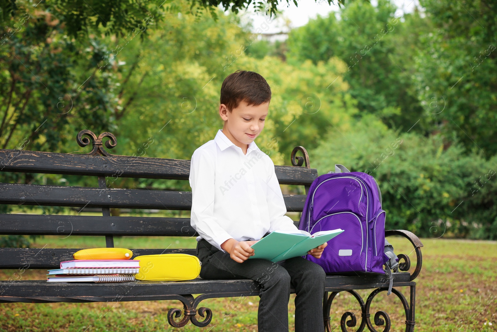 Photo of Cute little school child with stationery reading book on bench in park