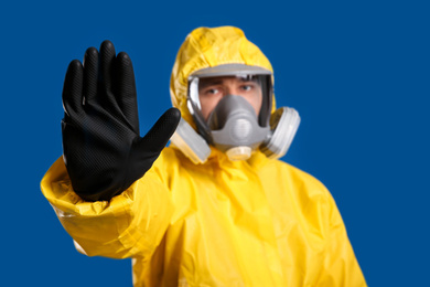 Photo of Man in chemical protective suit making stop gesture against blue background, focus on hand. Virus research