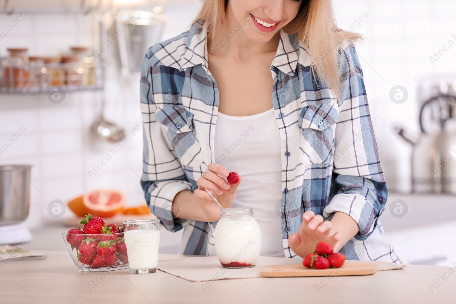 Photo of Young woman adding strawberry into jar with tasty yogurt at table