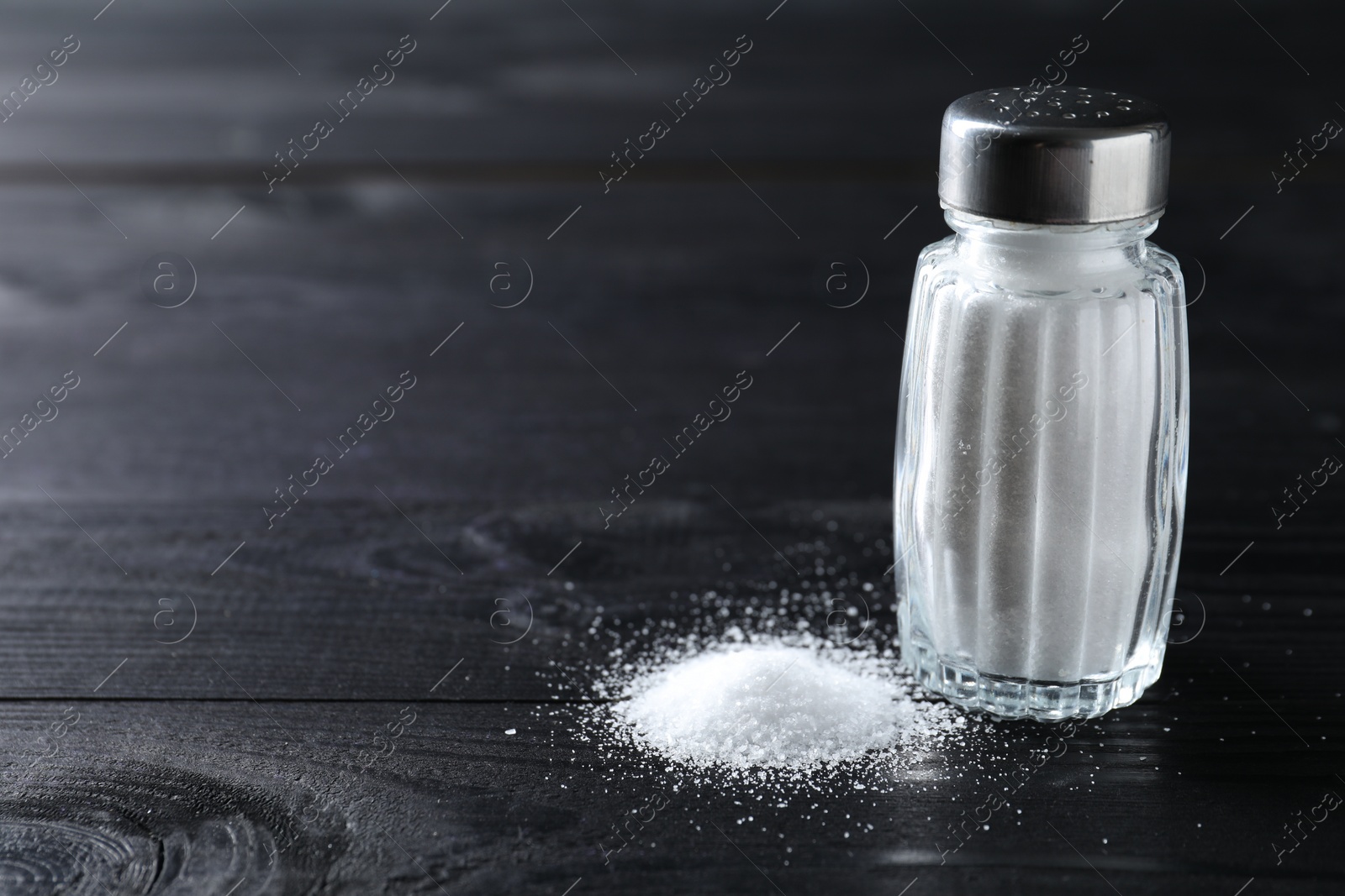 Photo of Organic salt in glass shaker on black wooden table, closeup. Space for text