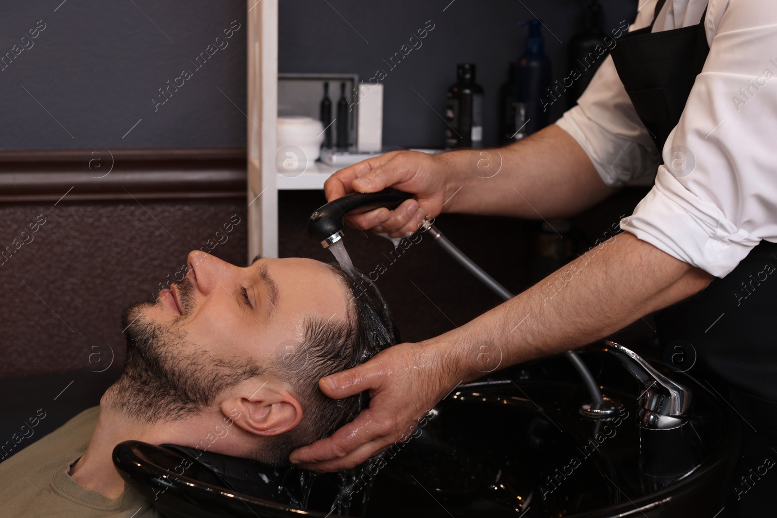 Photo of Professional hairdresser washing man's hair at sink in barbershop