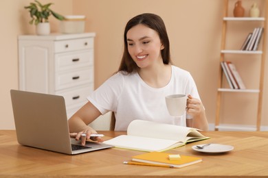 Photo of Happy young woman with cup of coffee and notebook working on laptop at wooden table indoors