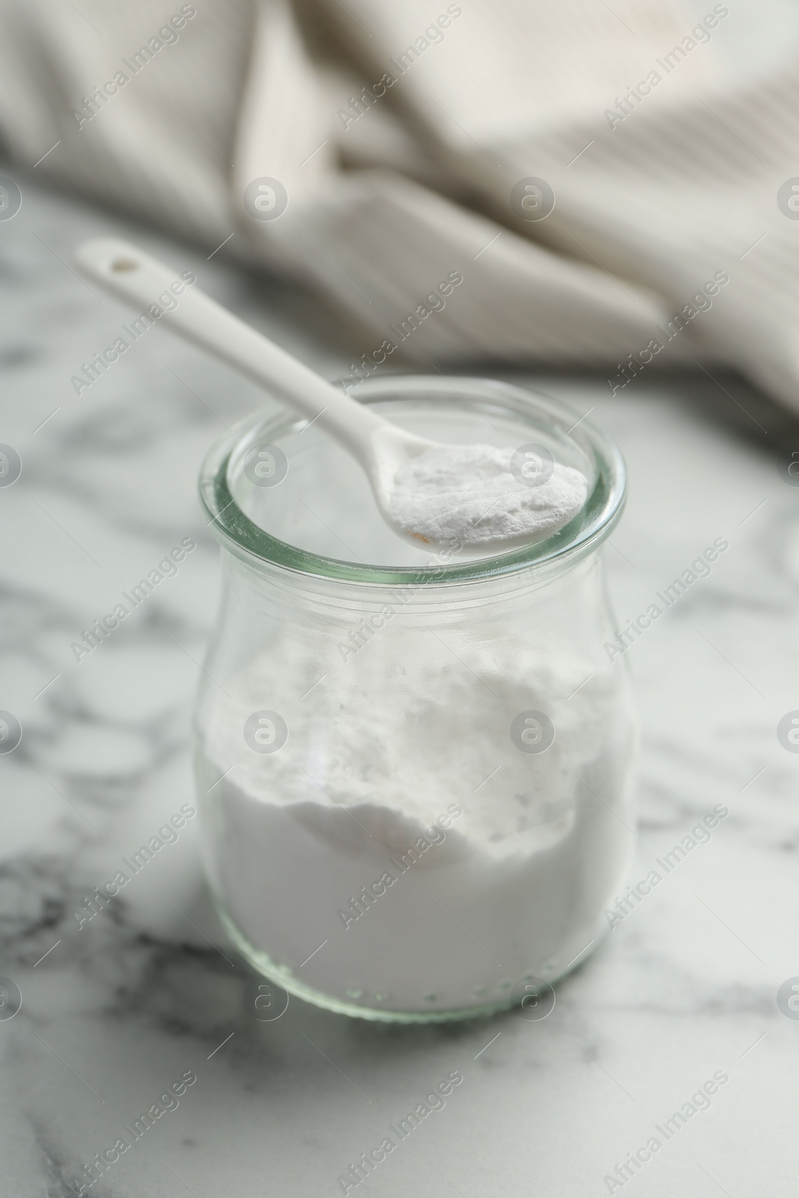 Photo of Baking powder in jar and spoon on white marble table, closeup