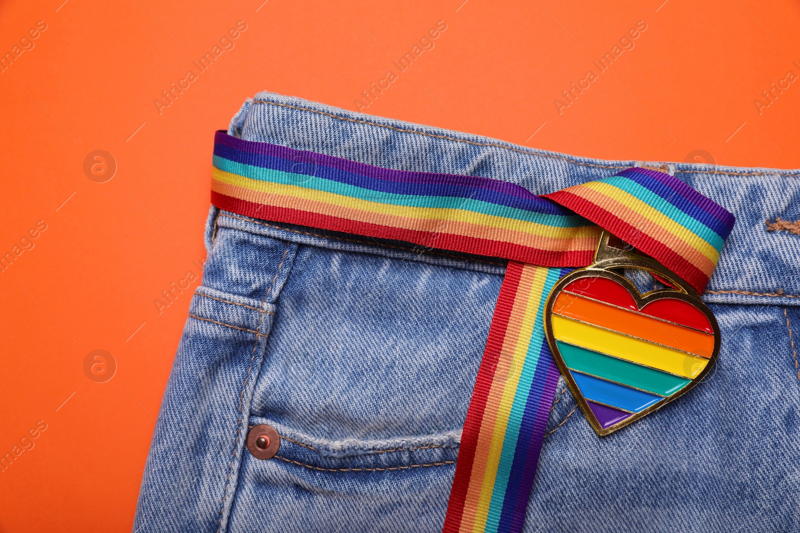 Photo of Jeans and rainbow ribbon with heart shaped pendant on orange background, top view. LGBT pride