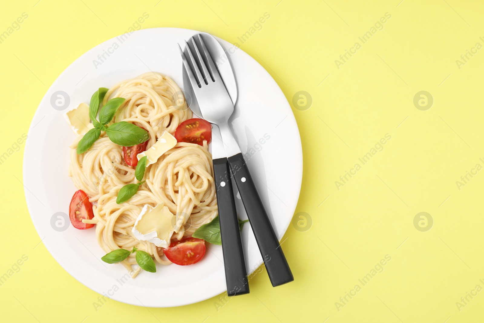 Photo of Delicious pasta with brie cheese, tomatoes, basil and cutlery on yellow background, top view. Space for text