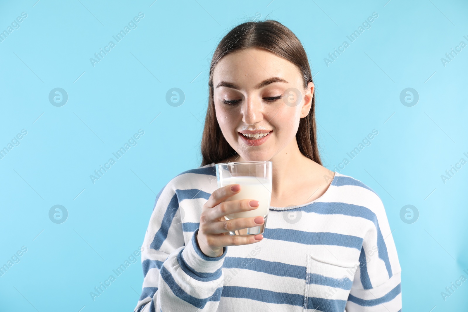 Photo of Beautiful woman with milk mustache holding glass of tasty dairy drink on light blue background