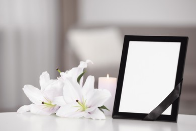 Image of Funeral photo frame with black ribbon, white and violet lilies on white table indoors. Space for text