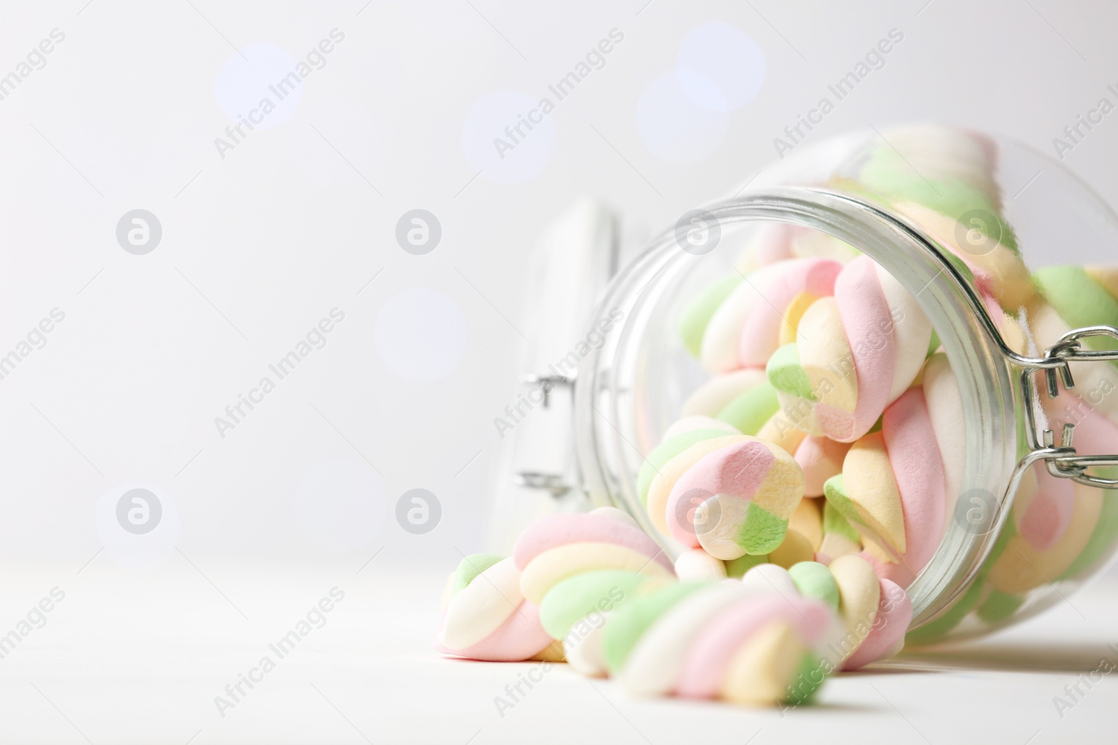 Photo of Glass jar with colorful marshmallows on white table against blurred lights, closeup. Space for text