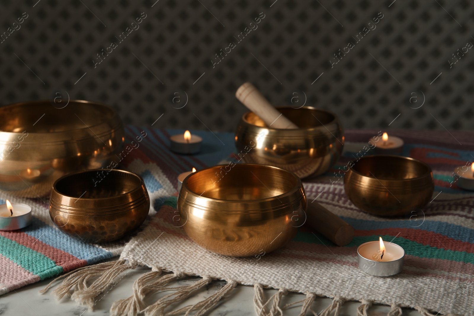 Photo of Tibetan singing bowls with mallets, burning candles and colorful fabric on white table