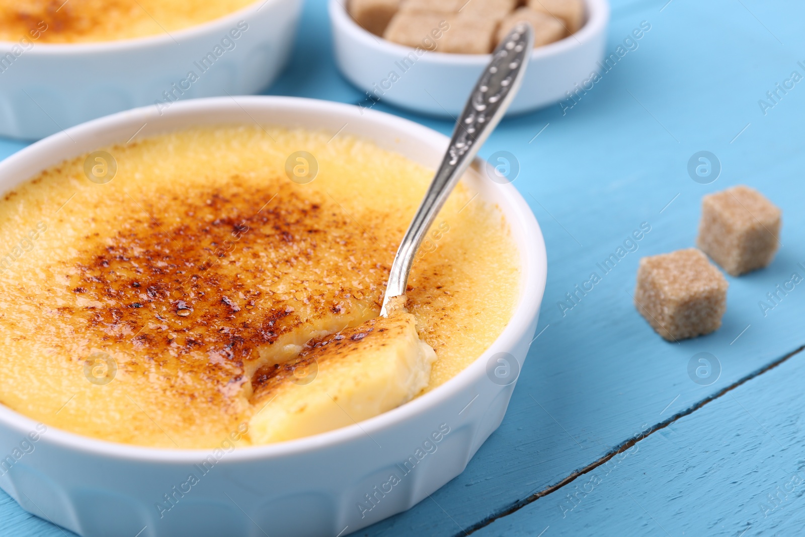 Photo of Delicious creme brulee in bowl, sugar cubes and spoon on light blue wooden table, closeup