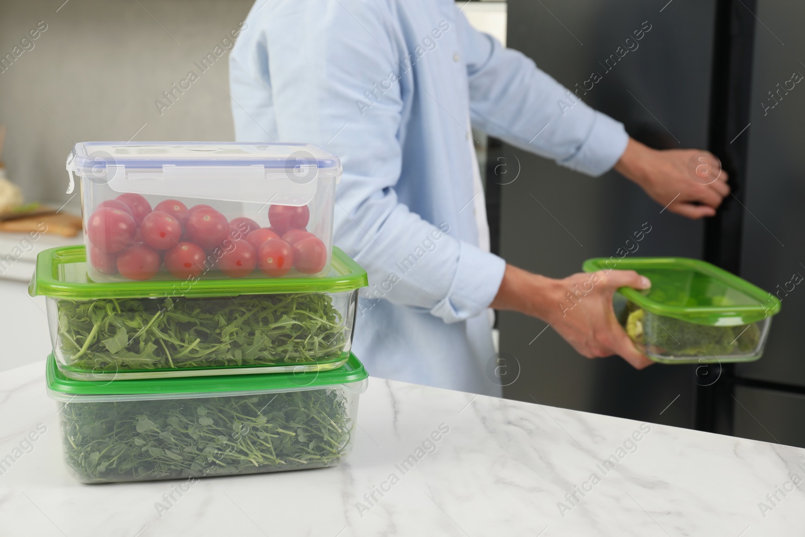 Photo of Glass and plastic containers with different fresh products on white marble table, selective focus. Man near fridge in kitchen