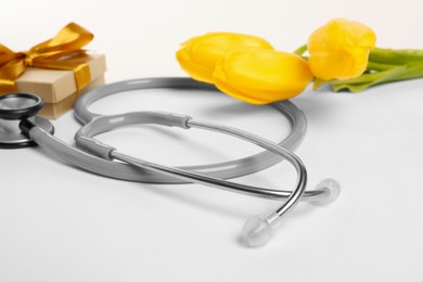 Photo of Stethoscope, gift box and yellow tulips on white background, closeup. Happy Doctor's Day