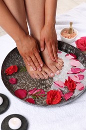 Photo of Woman soaking her feet in plate with water and red rose petals on white towel, closeup. Pedicure procedure