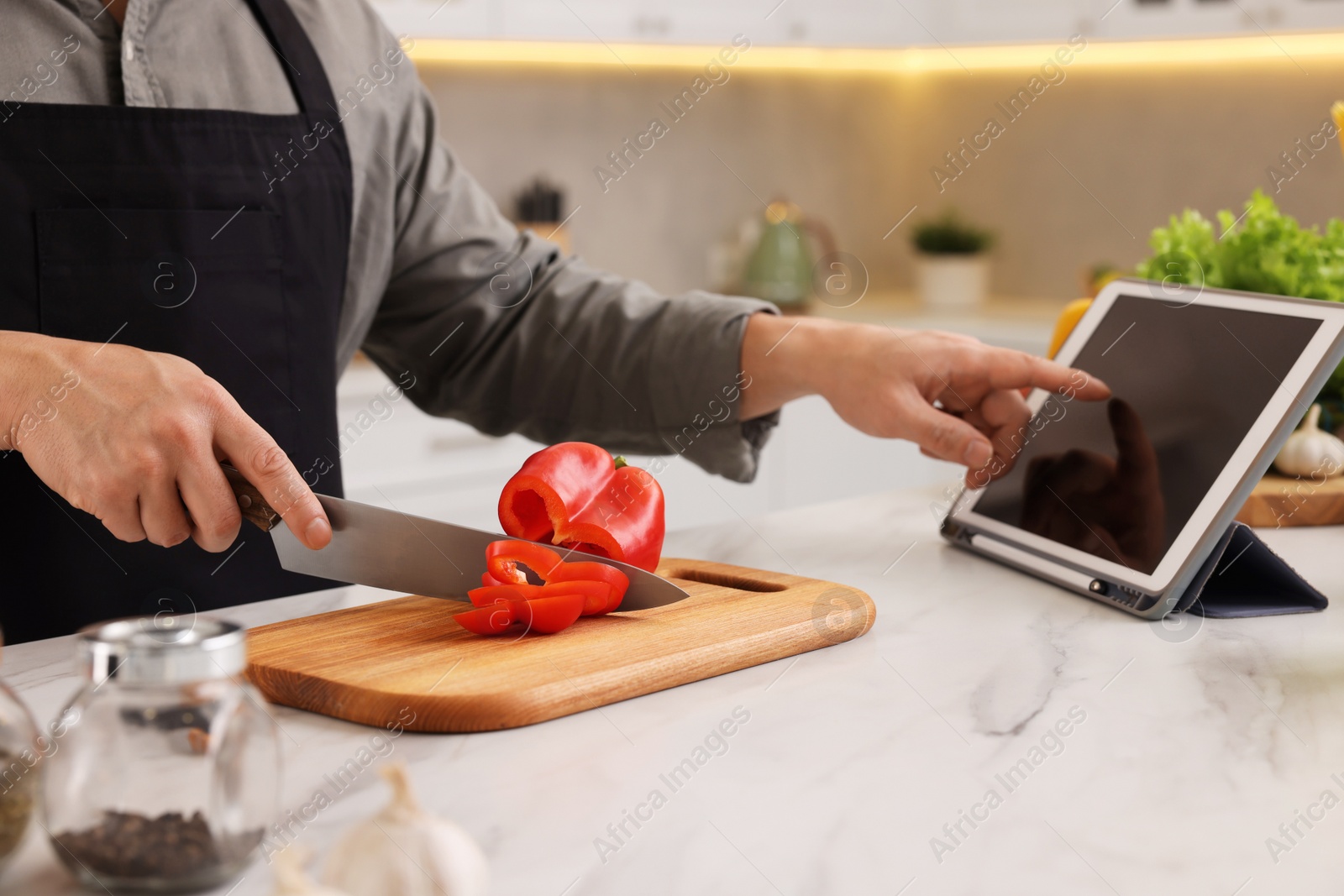 Photo of Cooking process. Man using tablet while cutting fresh bell pepper at white marble countertop in kitchen, closeup