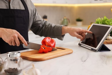 Photo of Cooking process. Man using tablet while cutting fresh bell pepper at white marble countertop in kitchen, closeup