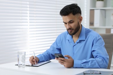 Handsome young man using smartphone while working at white table in office