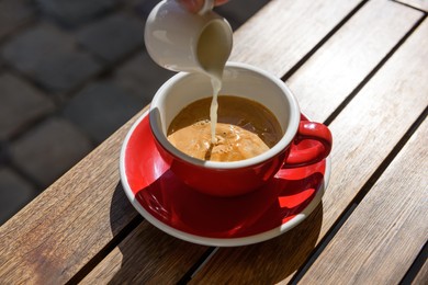 Pouring milk into cup of aromatic hot coffee at wooden table outdoors