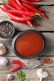 Photo of Spicy chili sauce in bowl and ingredients on wooden table, flat lay